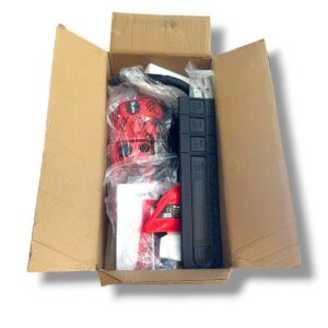 Open shipping box containing a red power tool, black storage module, and assorted accessories in plastic bags.