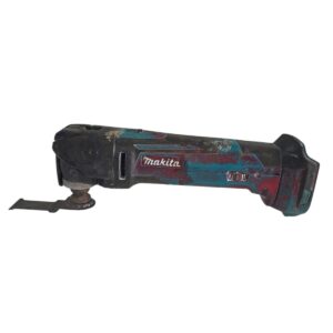 Cordless Makita oscillating multitool covered in dirt with blade attached, lying on its side on a white background
